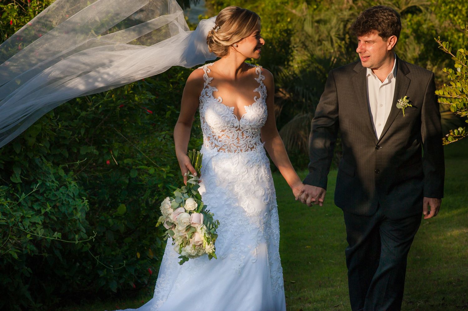 Bride in lace wedding dress and groom in tuxedo walking outdoors in golden light.