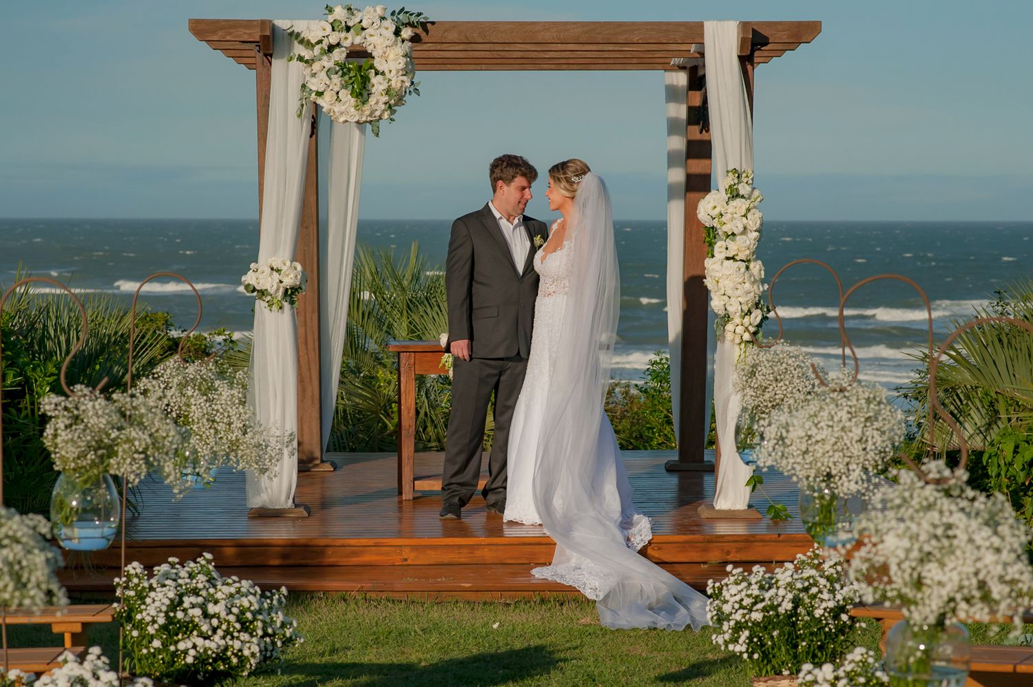 Wedding ceremony at wooden arch decorated with white flowers overlooking the ocean.