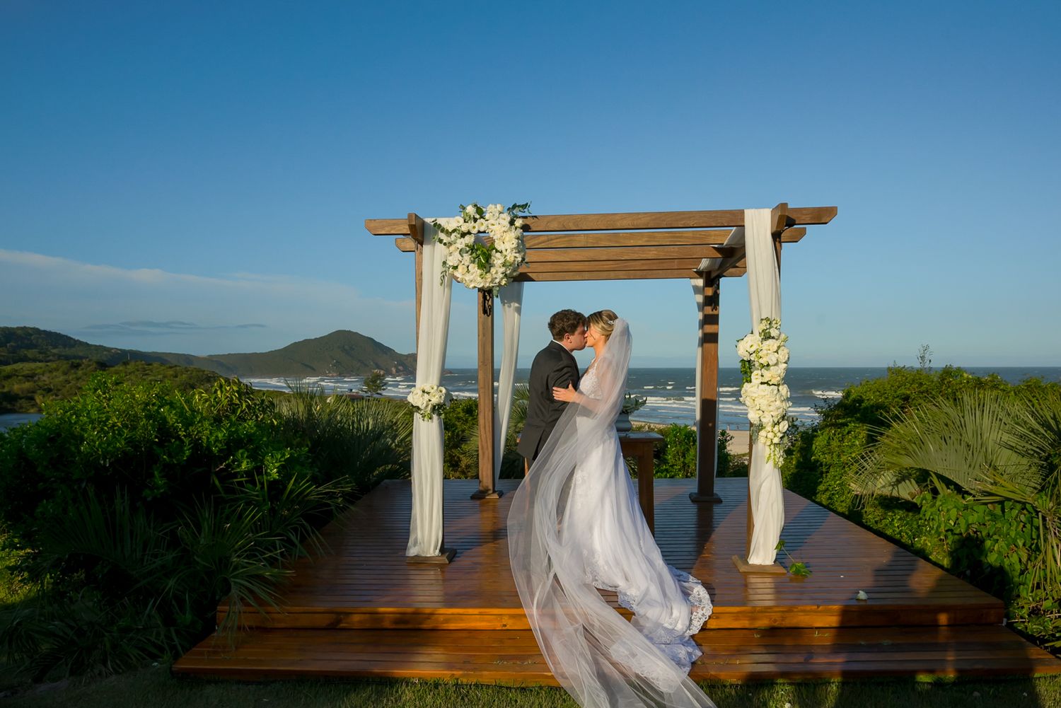 A scenic wedding ceremony setup with a wooden pergola decorated with white flowers and fabric overlooking mountains.