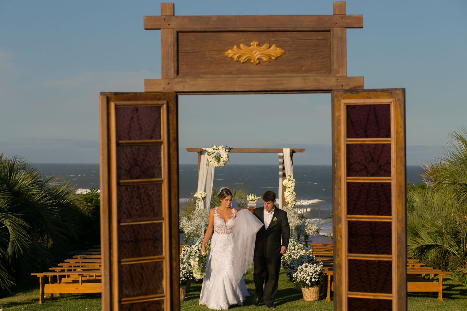 A rustic wooden wedding arch with large open doors framing an outdoor ceremony space with ocean views.