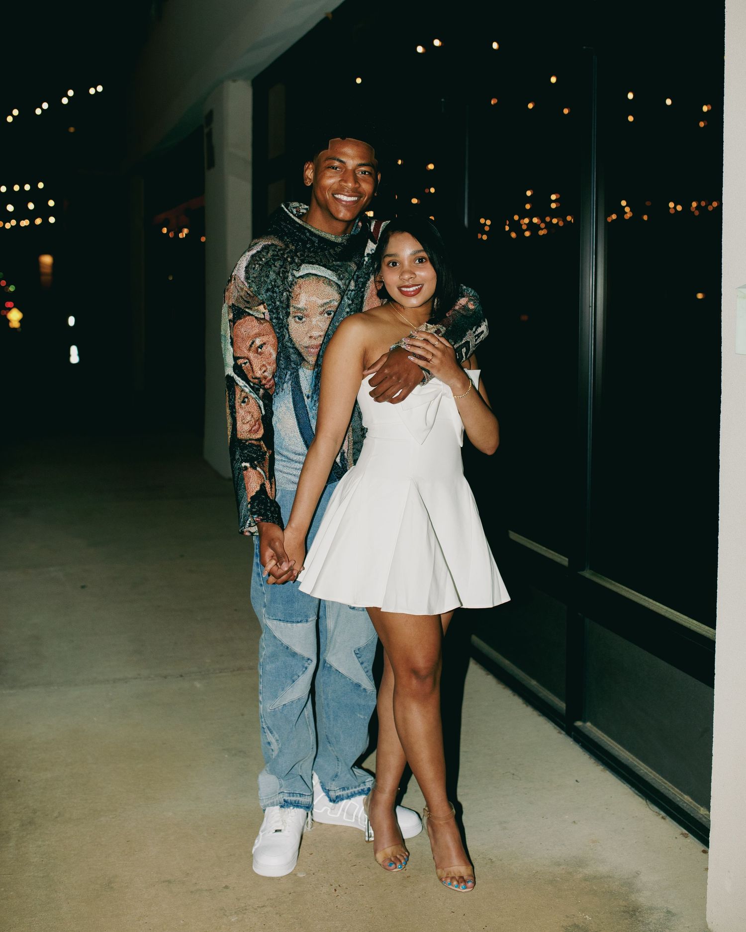 Couple in casual attire posing together in an outdoor setting with string lights in the background.