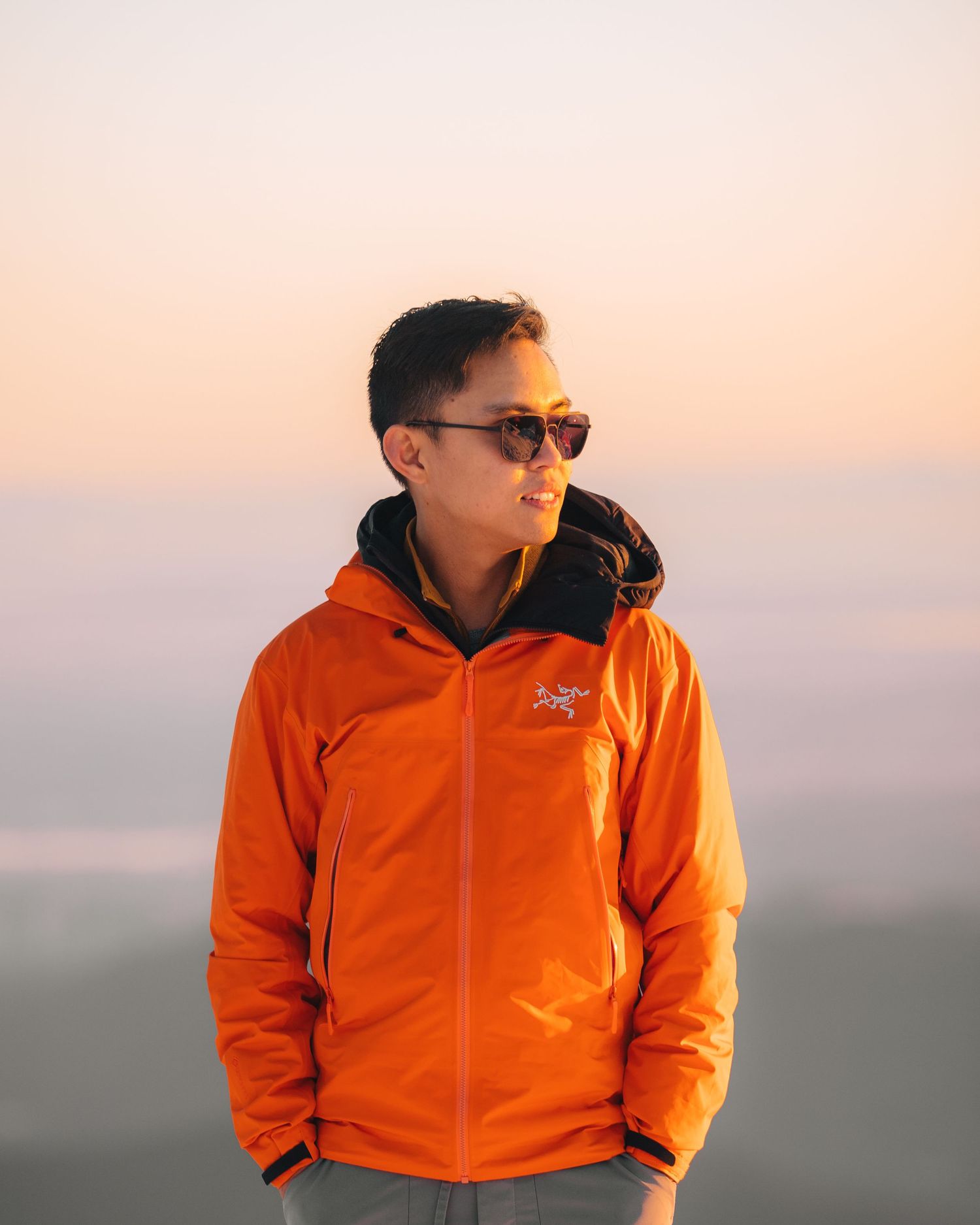 Person in bright orange jacket standing against sunset sky wearing sunglasses.