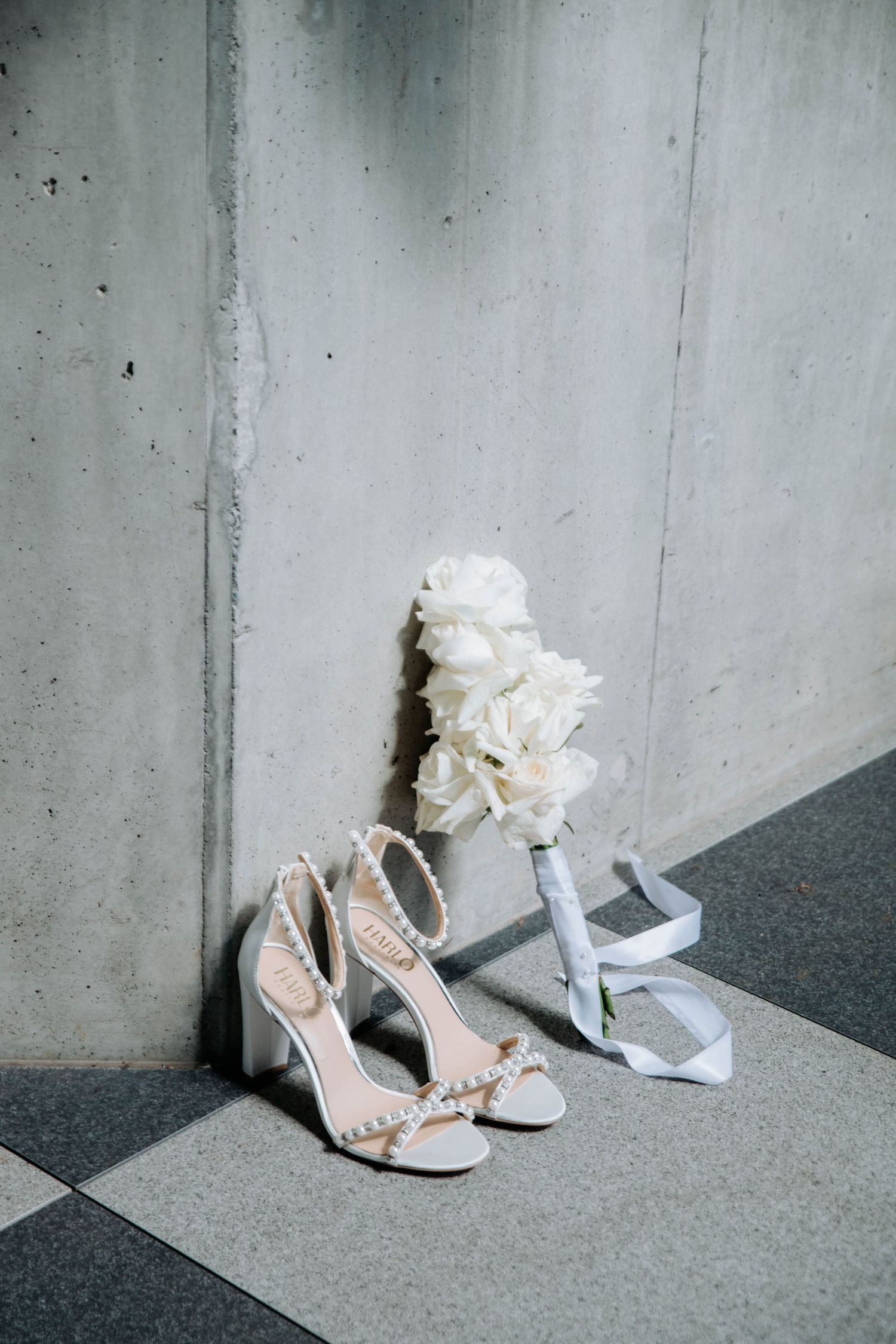 Pearl-embellished bridal shoes and white flowers against a concrete wall.