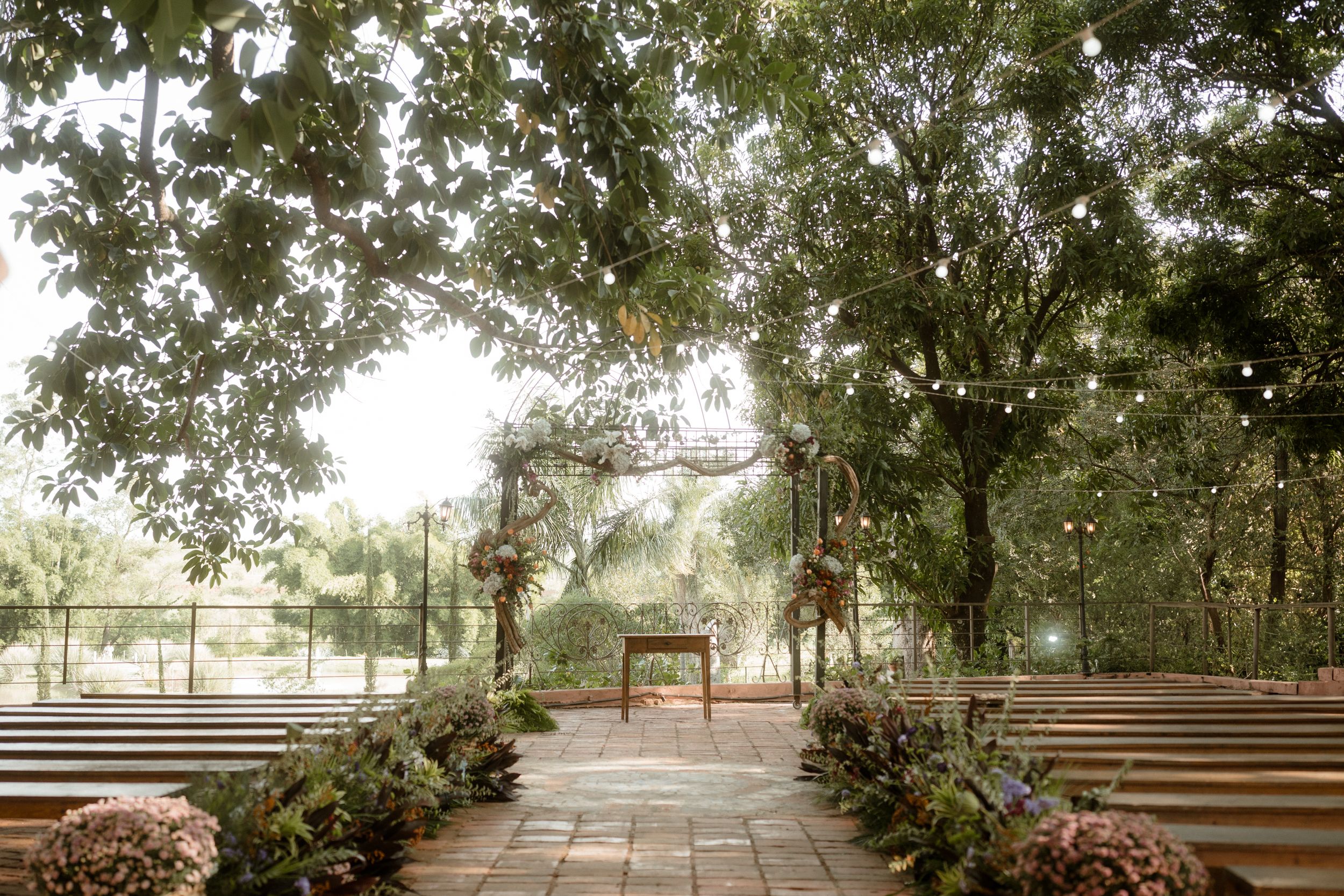 Rustic outdoor wedding ceremony setup with wooden benches arranged in rows under shady trees.