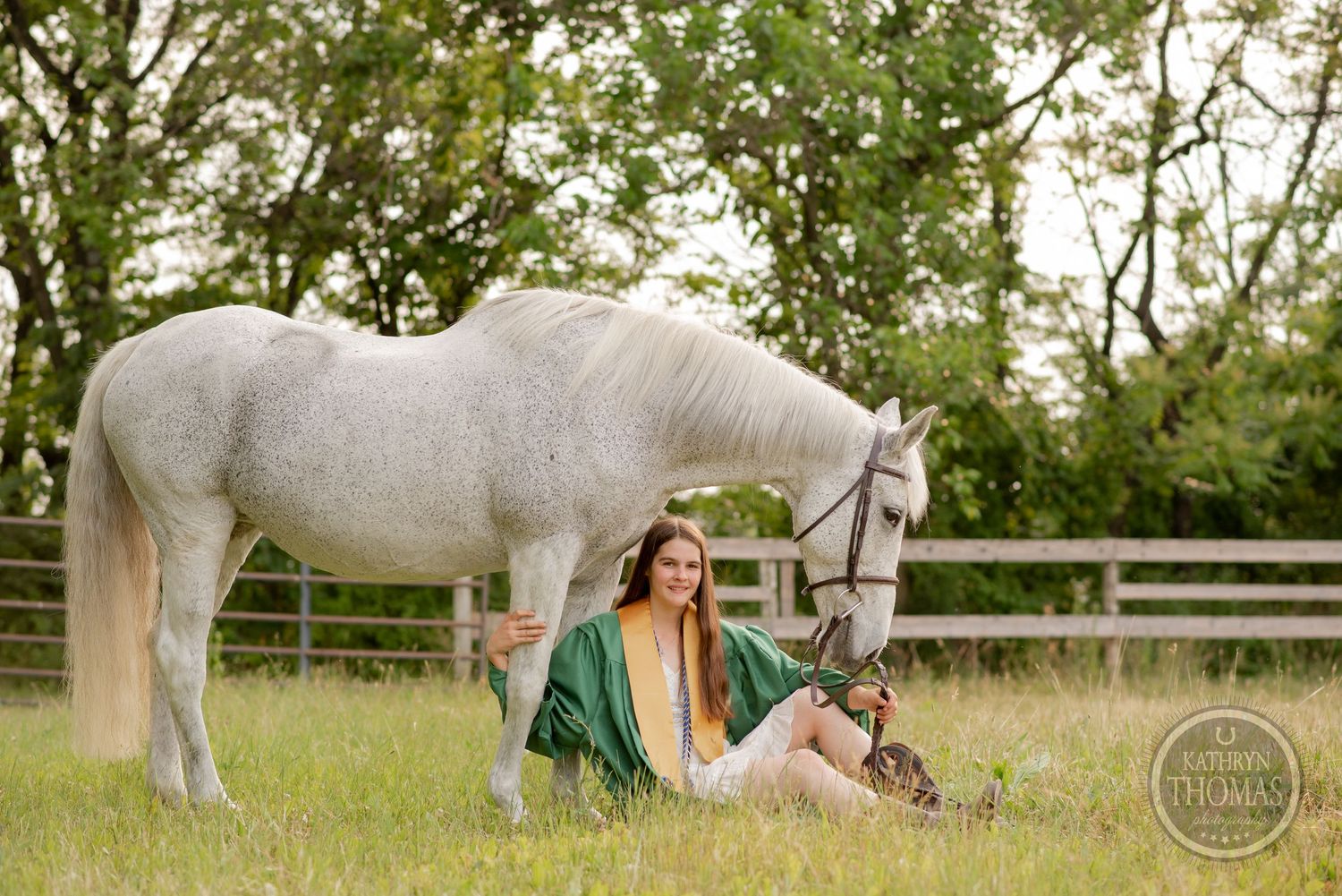 Graduate in green graduation robes relaxes in grass beside a white horse in pastoral setting.