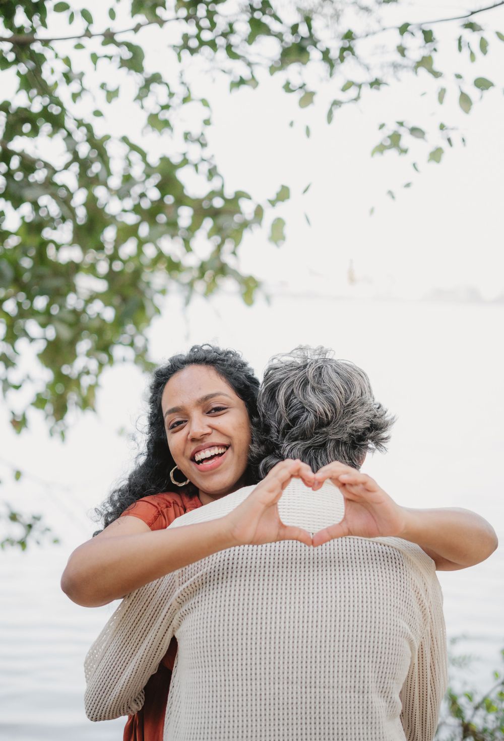 A person makes a heart shape with hands while embracing their partner under green trees.