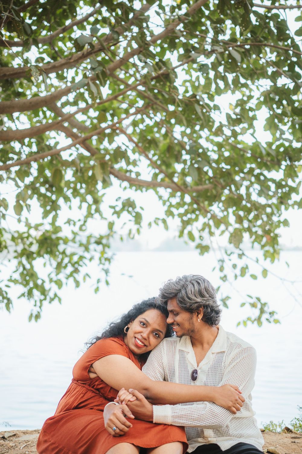 A couple embraces while sitting outdoors under a tree canopy near water.