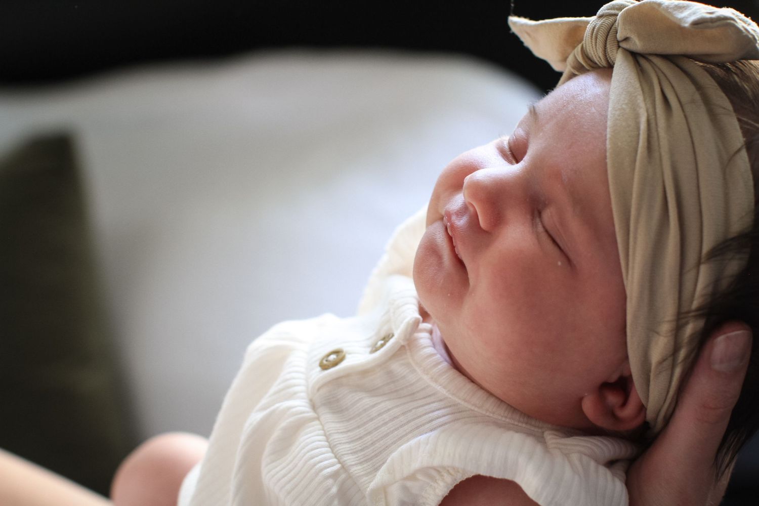 A closeup profile of a newborn baby girl in a bow headband sleeping and smiling peacefully in her father's hands.