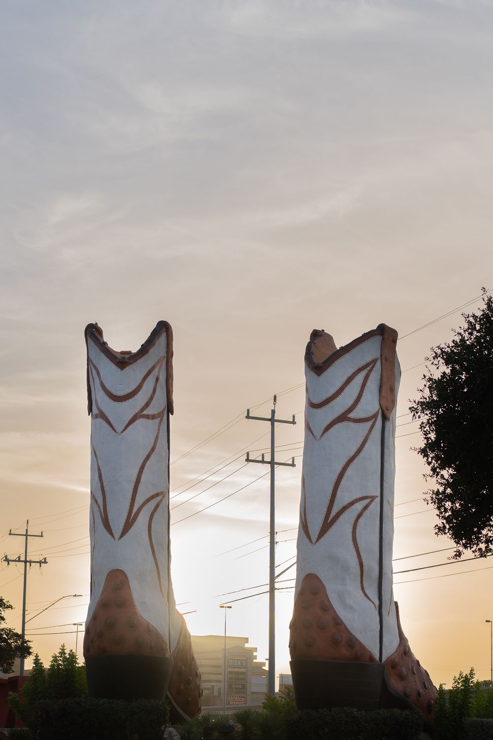 Giant decorative cowboy boots sculpture silhouetted against a sunset sky with power lines.