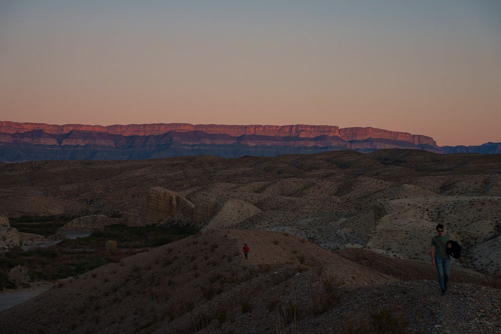 Desert landscape panorama at sunset with distant mountains and rocky terrain silhouetted against orange sky.