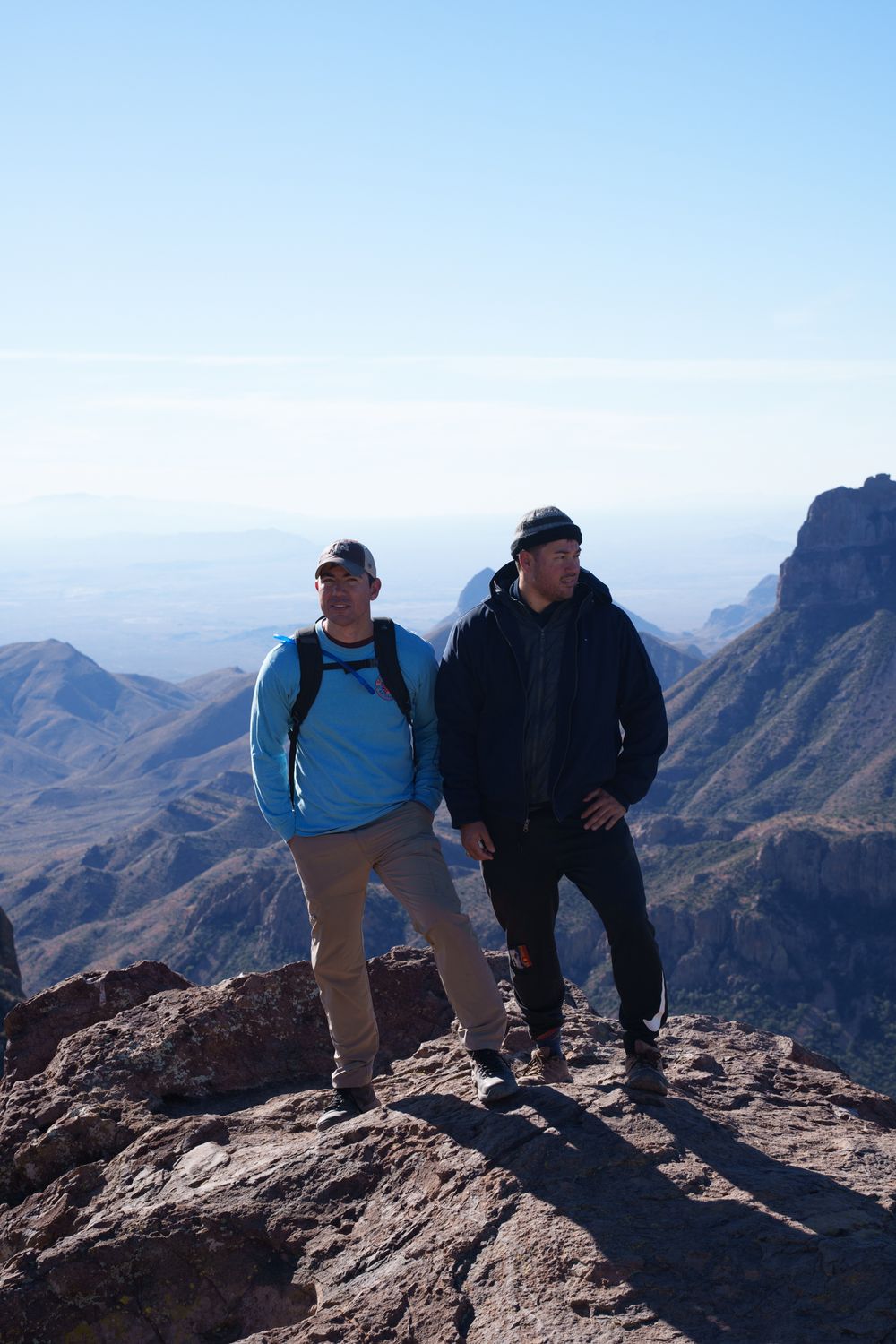 Two hikers stand at a scenic overlook on a rocky mountain peak with dramatic desert landscape views in the background.
