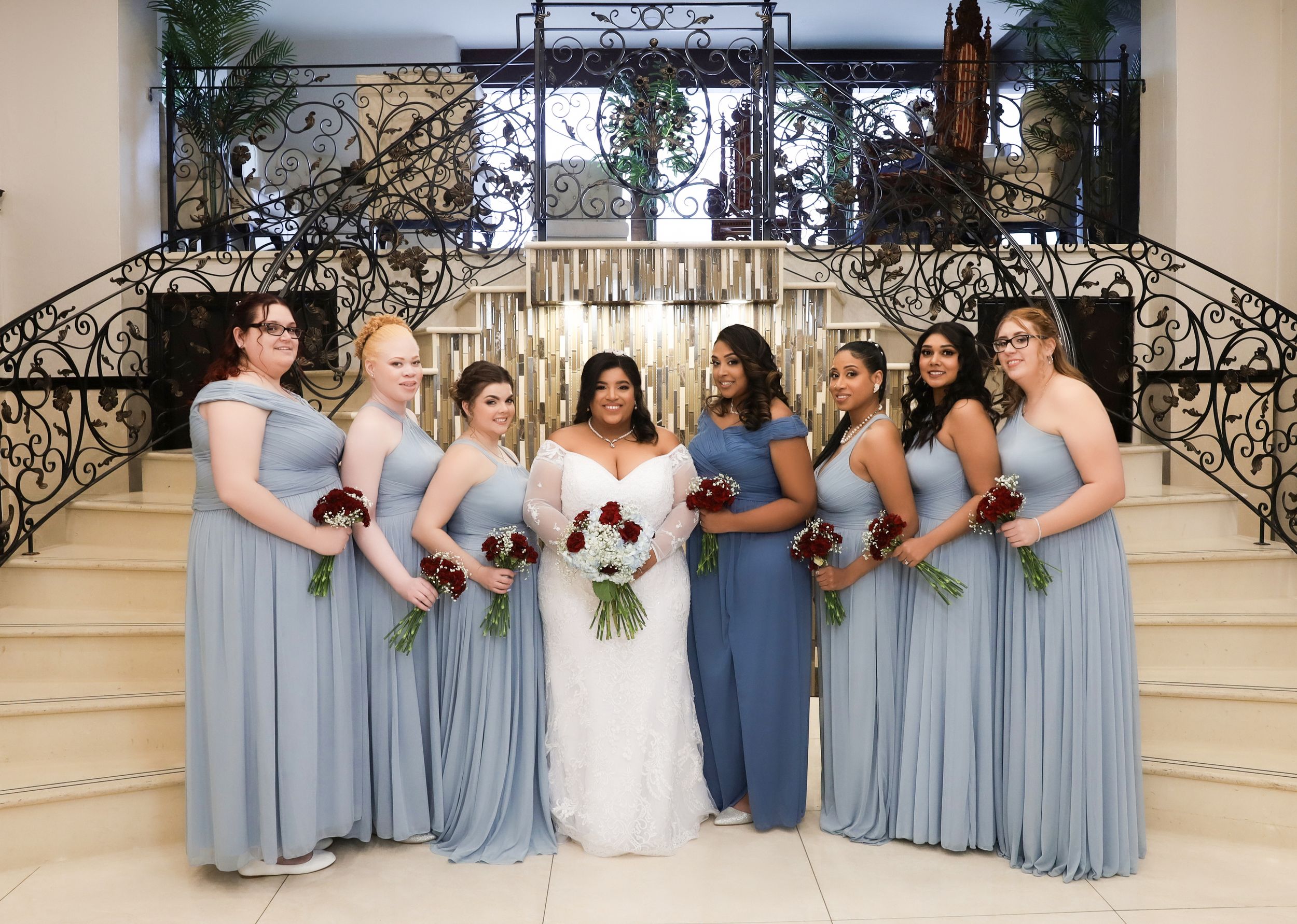 Bridal party in light blue dresses poses on grand staircase with ornate iron railings.