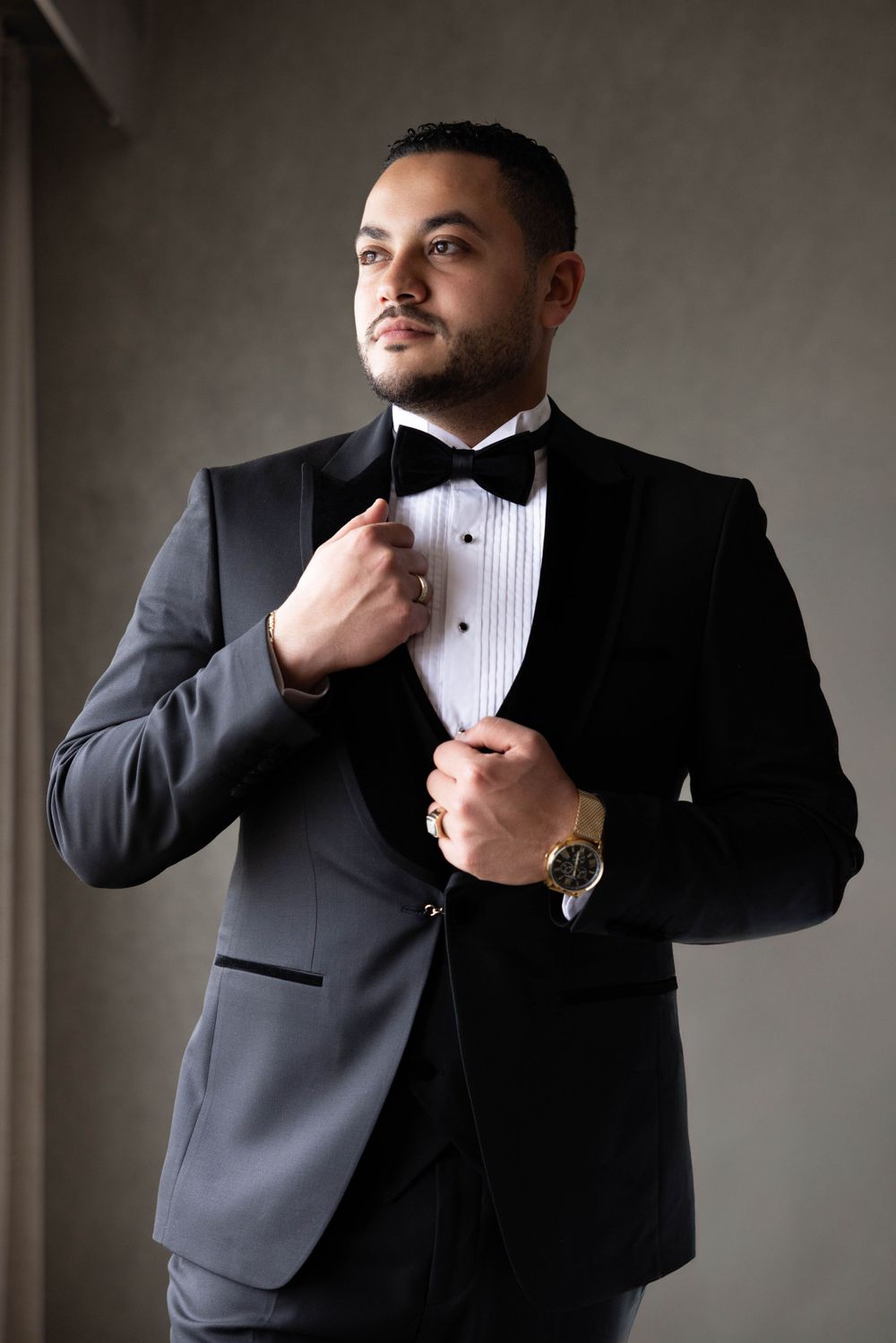 Groom adjusts his bowtie while wearing classic black tuxedo in formal portrait.