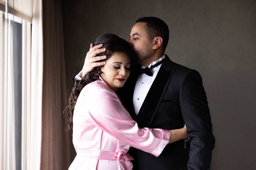Couple sharing an intimate moment by a window with person wearing pink jacket and formal black attire.