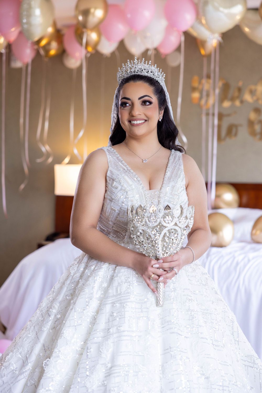 A bride in an elegant white lace ball gown and crystal tiara poses in front of pink and gold balloons at a celebration.