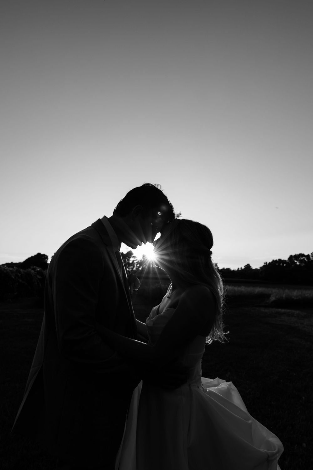 Dramatic black and white silhouettes of a wedding couple against sunset with light rays peeking through.