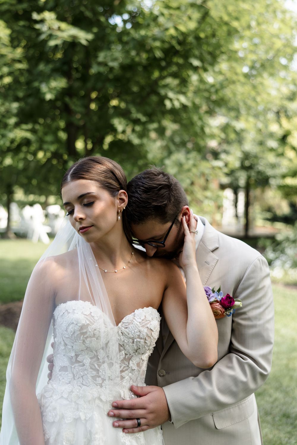 A couple embraces tenderly in an outdoor garden setting wearing elegant wedding attire.