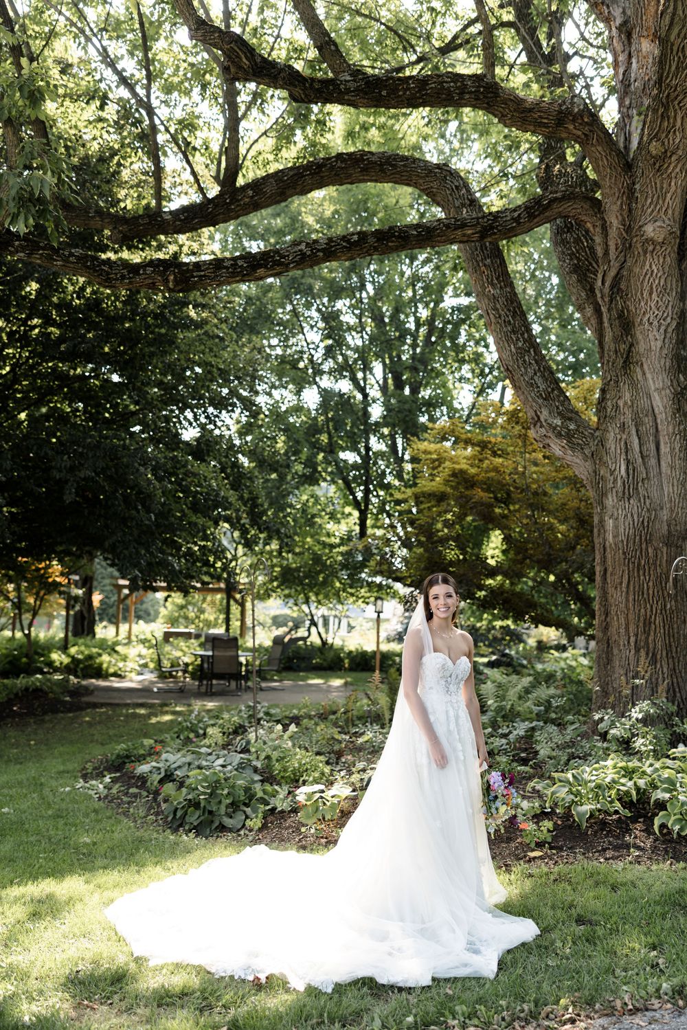 A white wedding dress trails elegantly across green grass beneath towering trees in a garden setting.