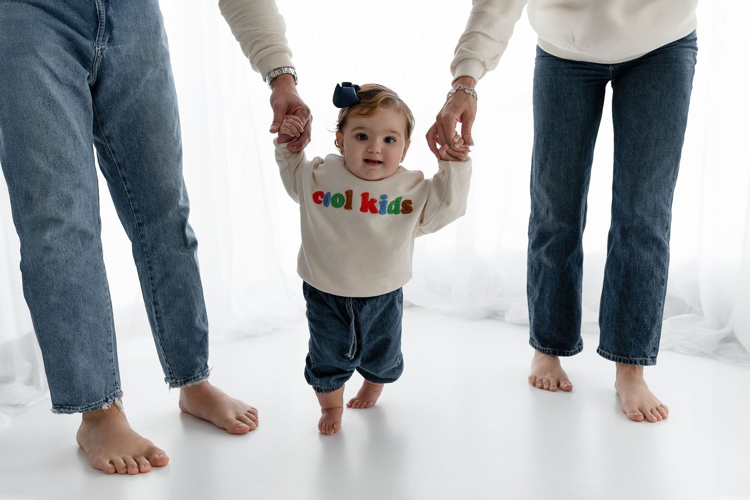 Small child takes first steps while holding parents hands against white backdrop.