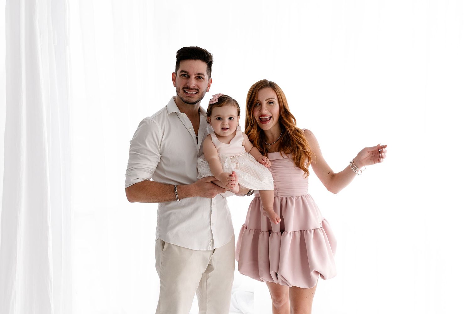 A happy family of three in light colored outfits poses together against a white backdrop.