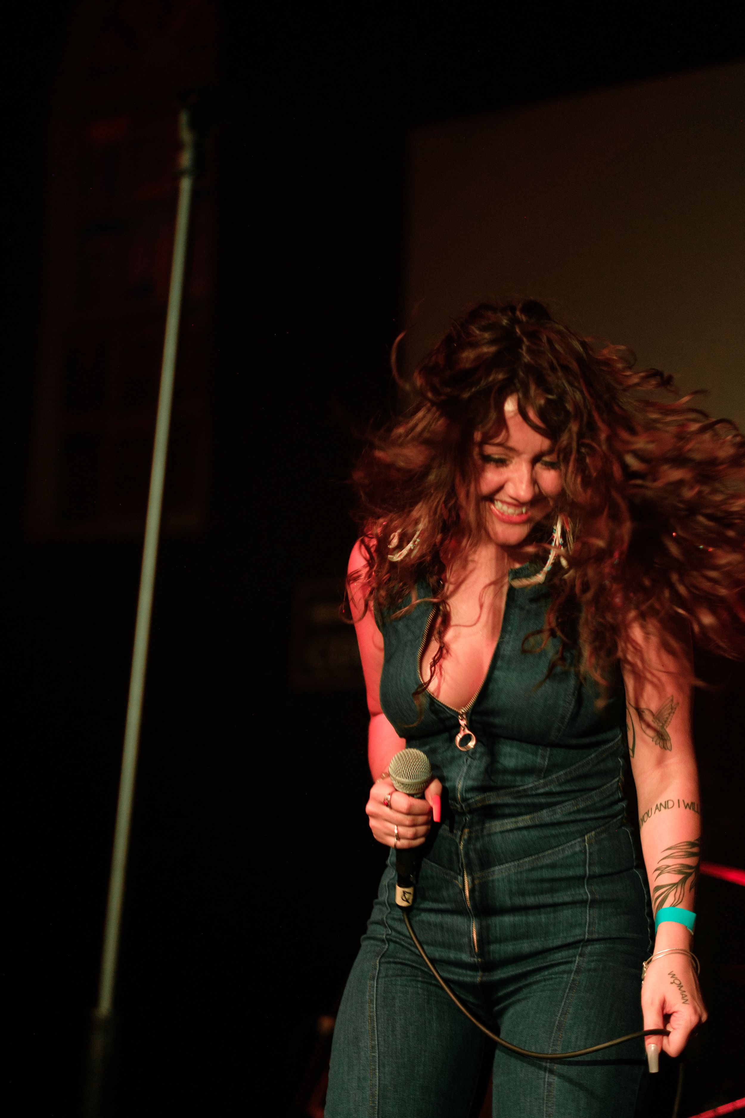Energetic performer with long flowing hair singing passionately on stage in dark lighting.