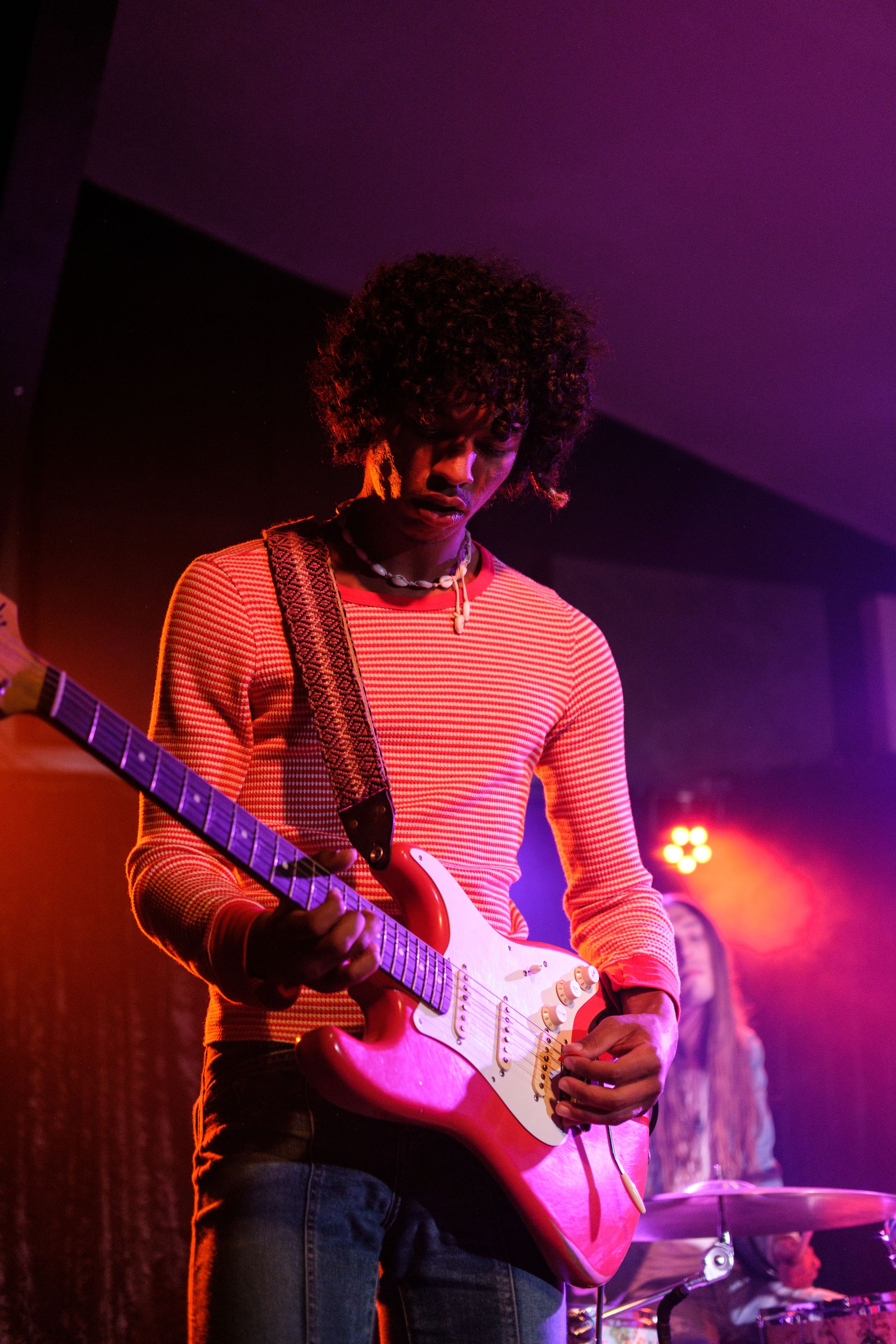 Guitarist in a striped shirt performs under purple stage lighting at a concert.