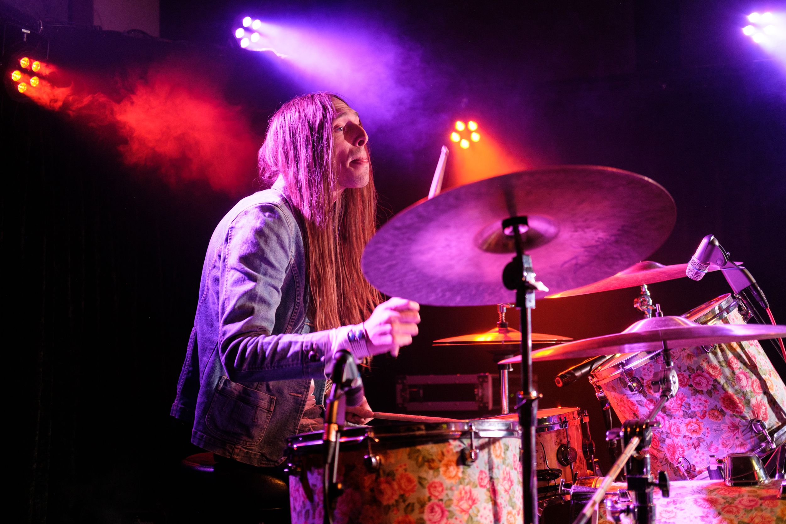 Drummer with long hair performing energetically behind a drum kit under colorful stage lights.