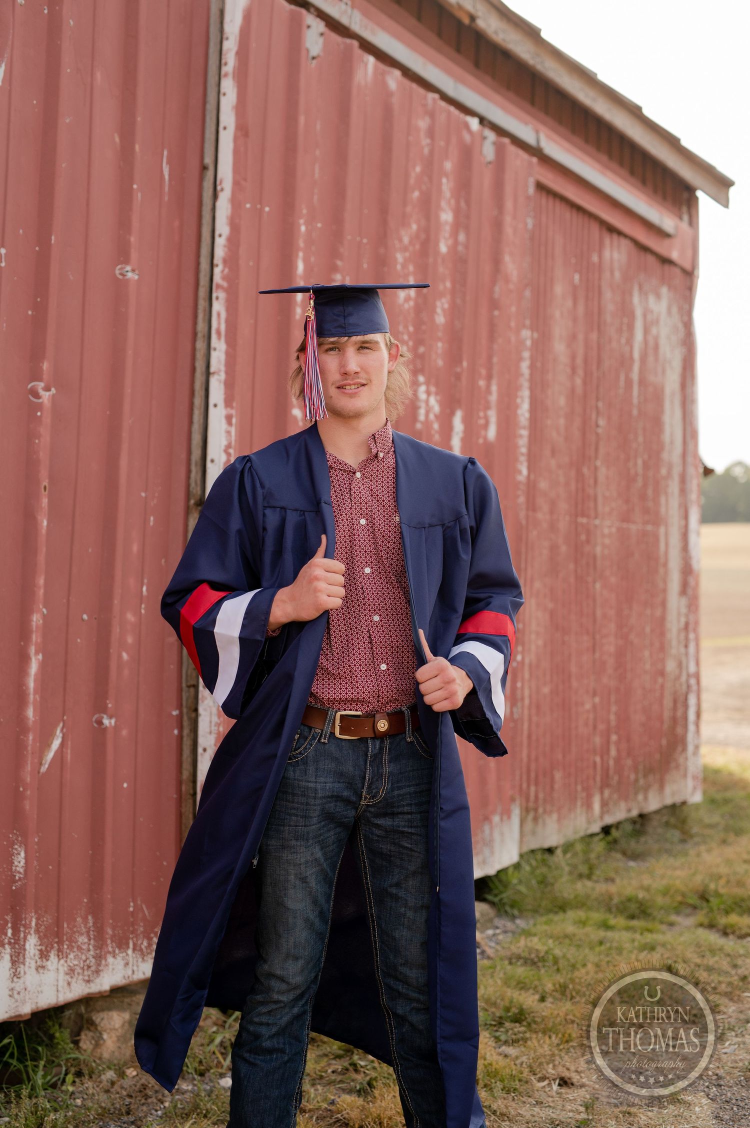 Graduate in navy gown with red and white stripes poses casually by red barn.