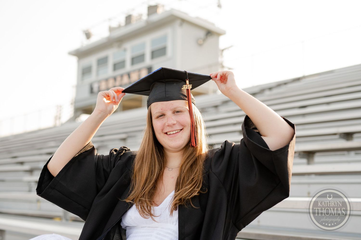 Graduate adjusts cap while smiling in front of stadium press box.