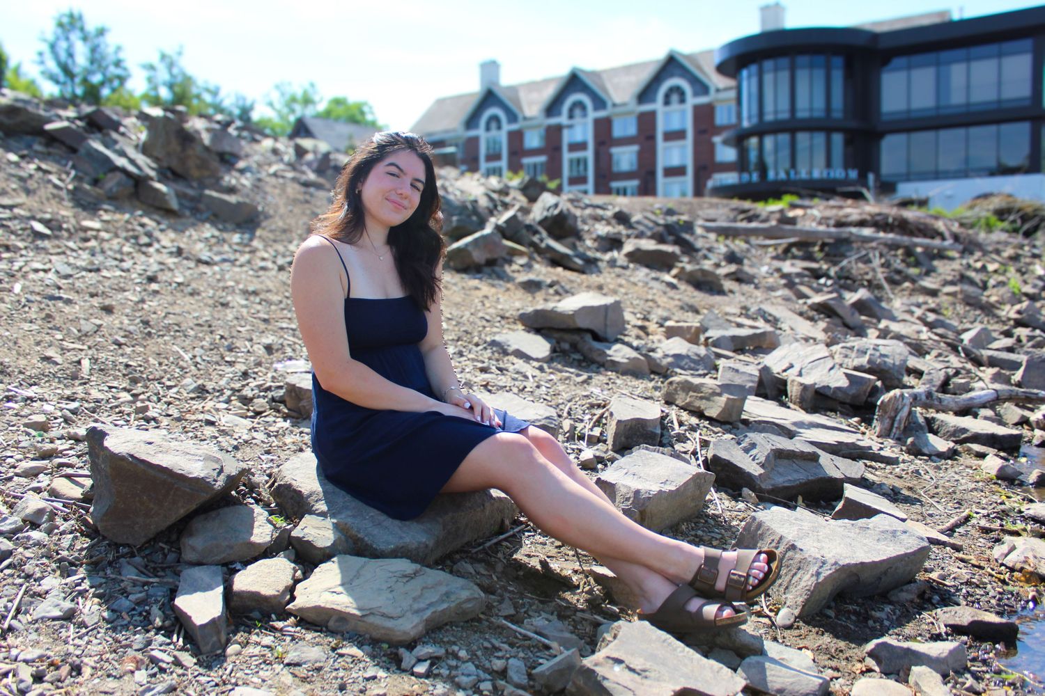 A figure in a navy dress sits on rocky terrain with buildings visible in the background.