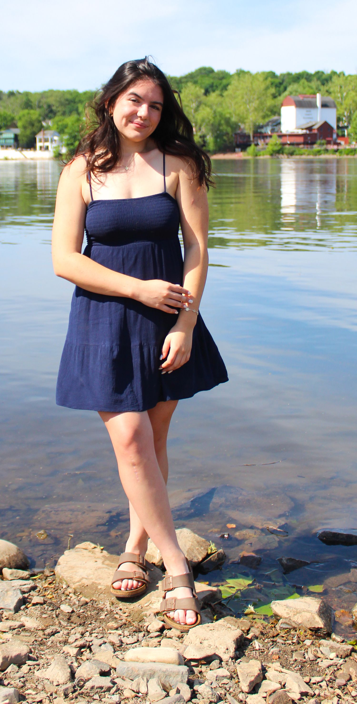 A figure in a navy blue sundress poses on rocks by a serene lakefront with houses in the distance.