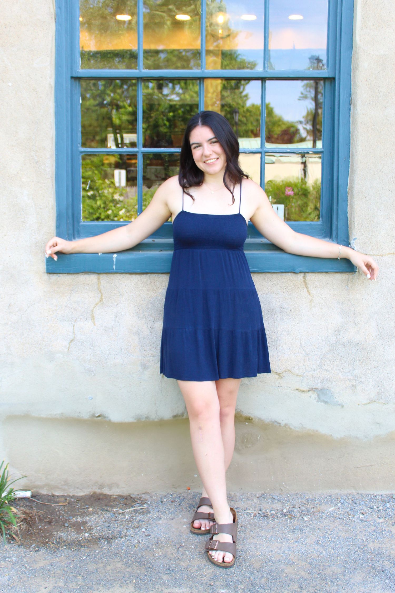 A figure in a navy dress poses against a white wall with a blue-trimmed window.