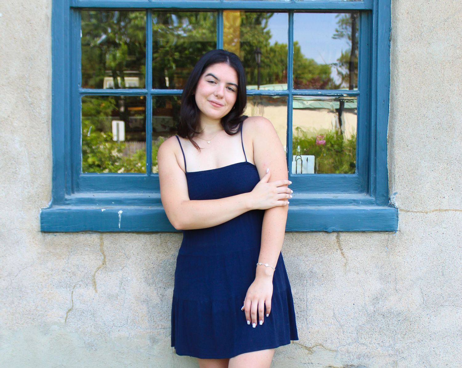A figure stands by a blue-trimmed window wearing a navy sundress against a white stucco wall.