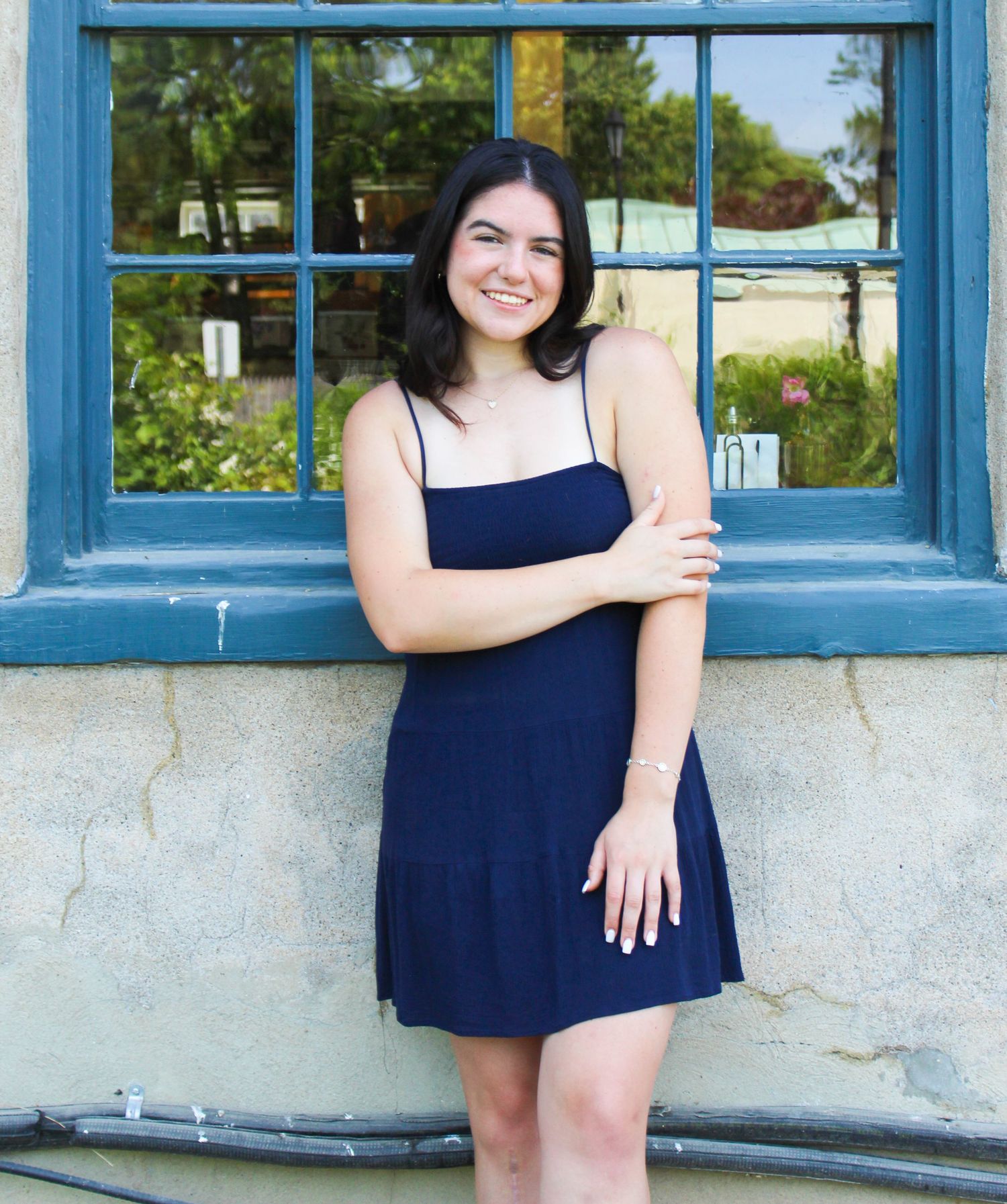 A figure poses in front of a blue window frame wearing a navy dress against a textured wall.