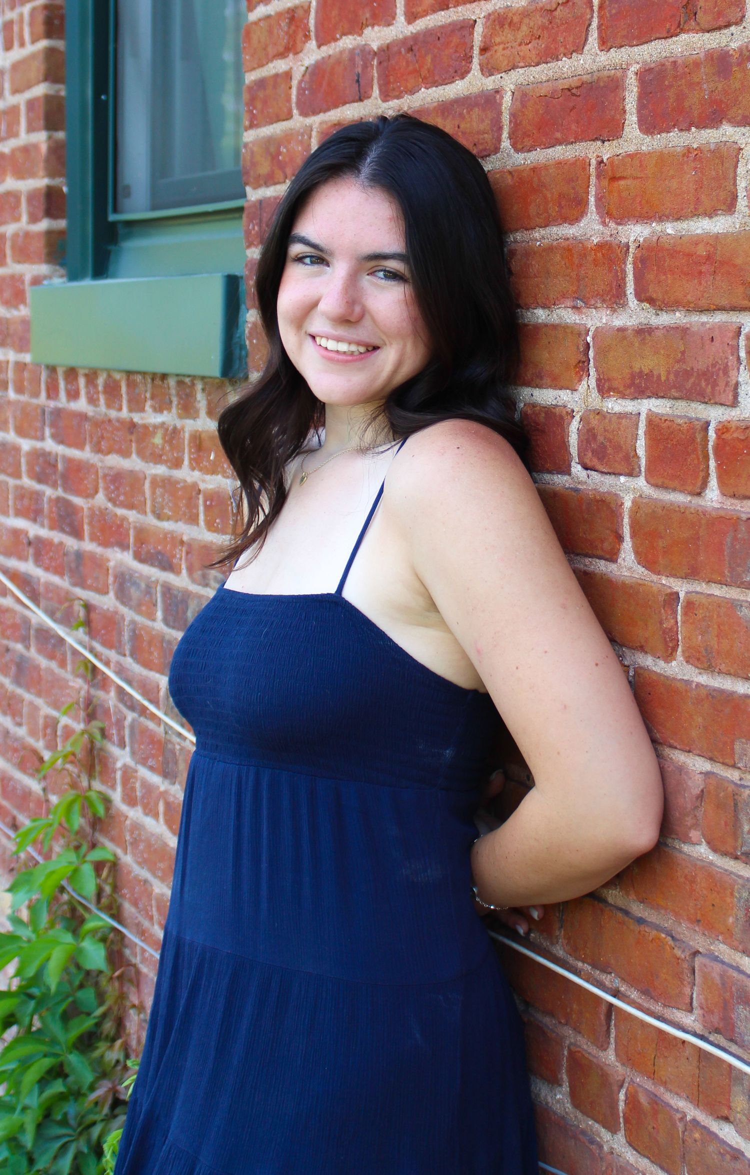 A figure in a navy dress poses against a red brick wall with a blue-trimmed window.