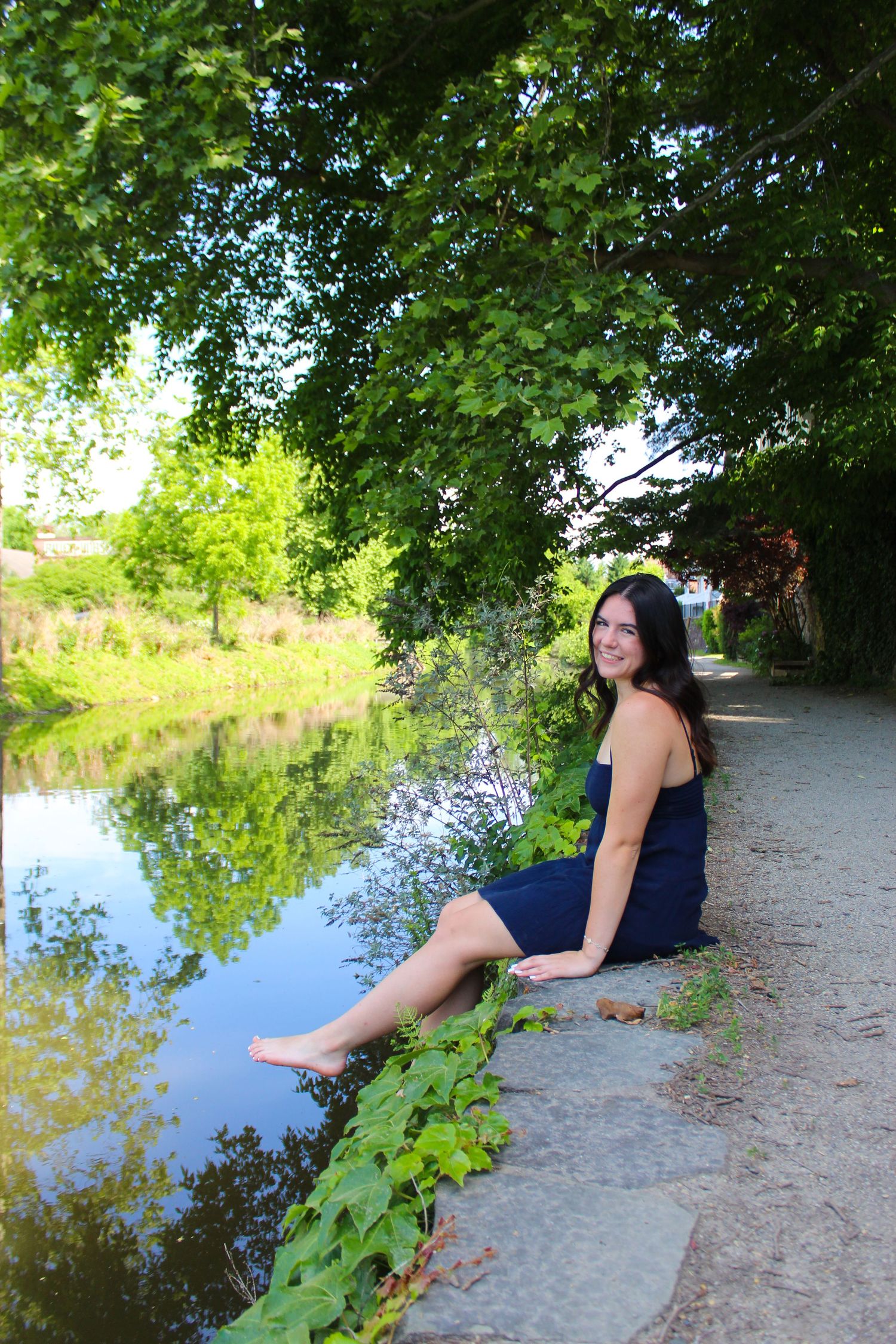 A figure in a navy dress sits along a scenic pathway next to a tranquil pond with overhanging trees.