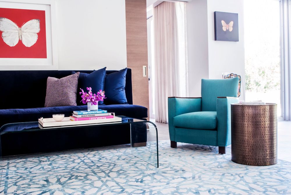 Modern living room with navy velvet sofa, teal armchair, butterfly art, and geometric blue rug.