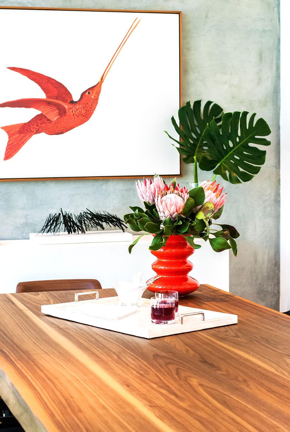 Modern tropical decor featuring red vase with protea flower, monstera leaf, and red hummingbird artwork on wooden table.