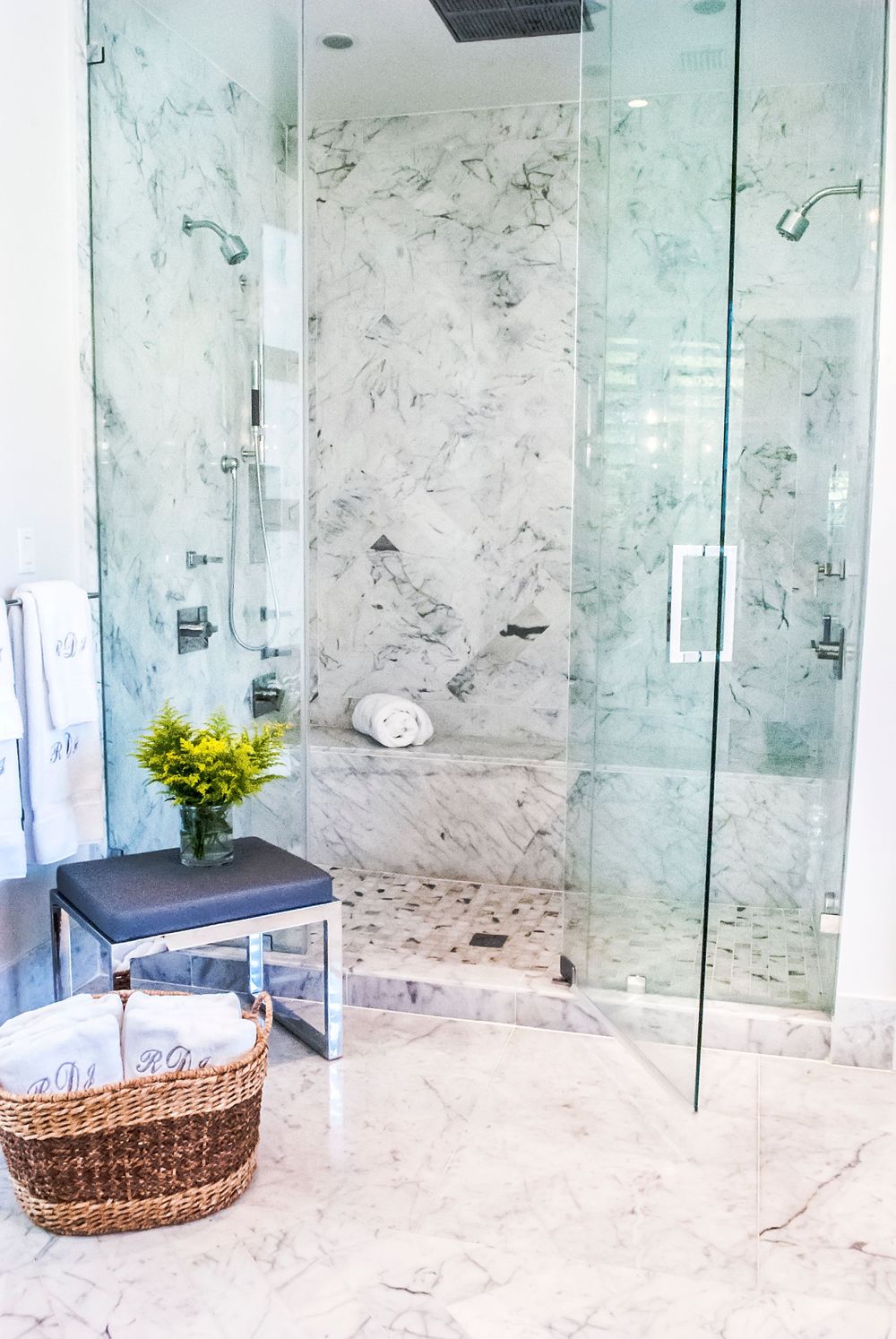 Modern marble bathroom interior featuring a glass shower enclosure, small bench with yellow flowers, and woven basket decor.