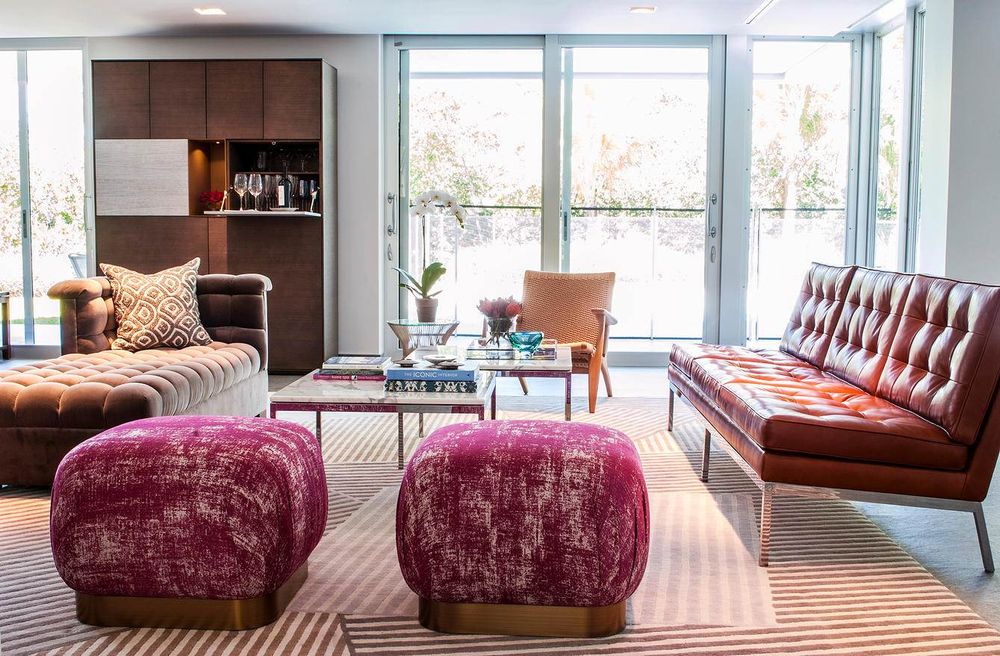 Contemporary living space featuring velvet burgundy poufs, leather seating, and floor-to-ceiling windows.