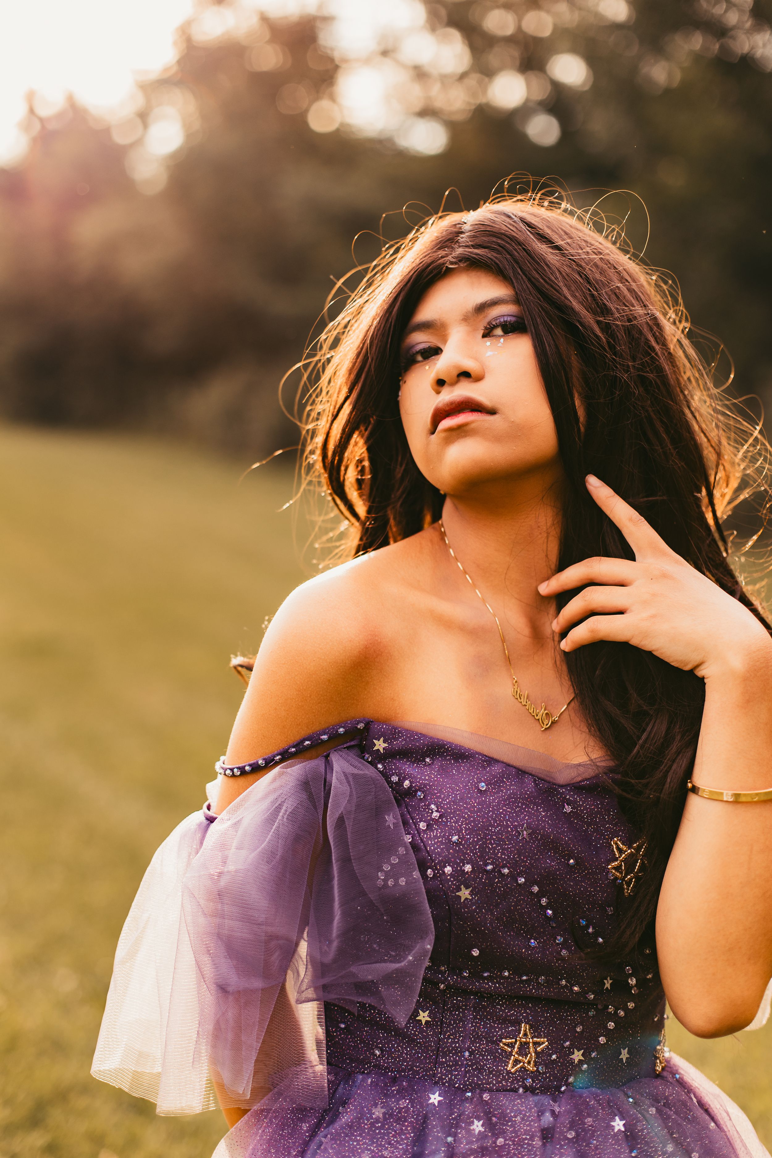 A person in a purple sparkly gown poses outdoors during golden hour with ethereal lighting and soft focus.