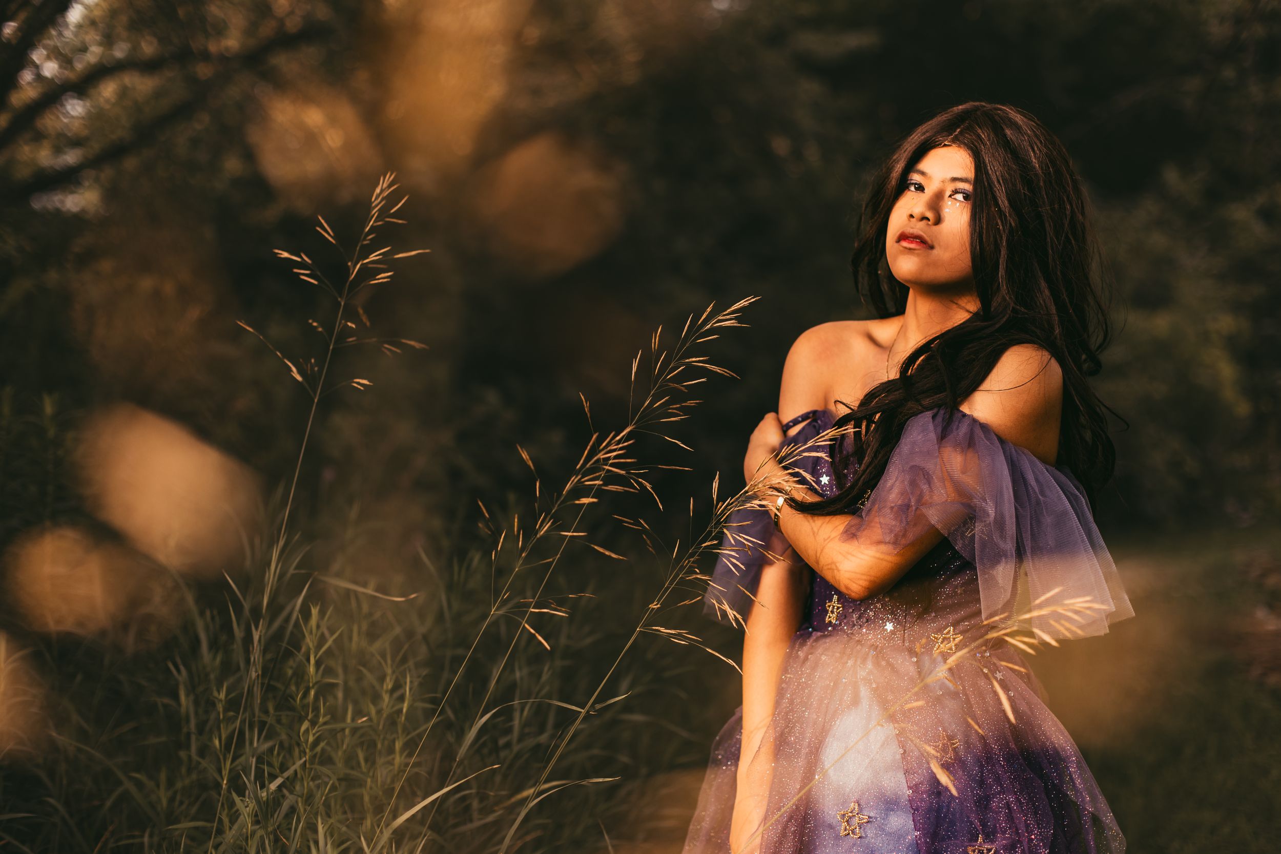 A woman in a flowing purple dress poses gracefully in a golden-lit field during sunset.