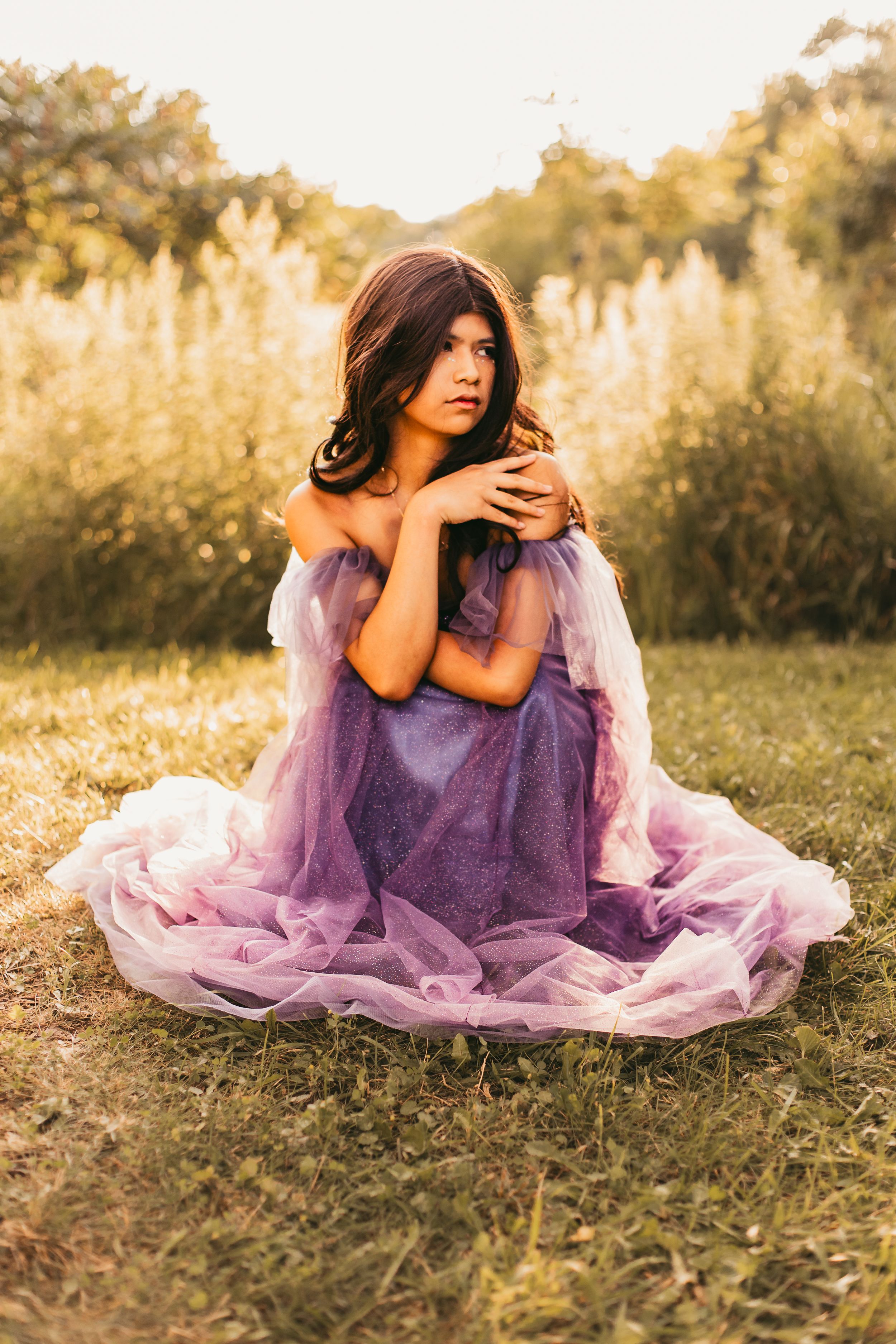 A person in a flowing purple dress sits gracefully on sunlit grass during golden hour.