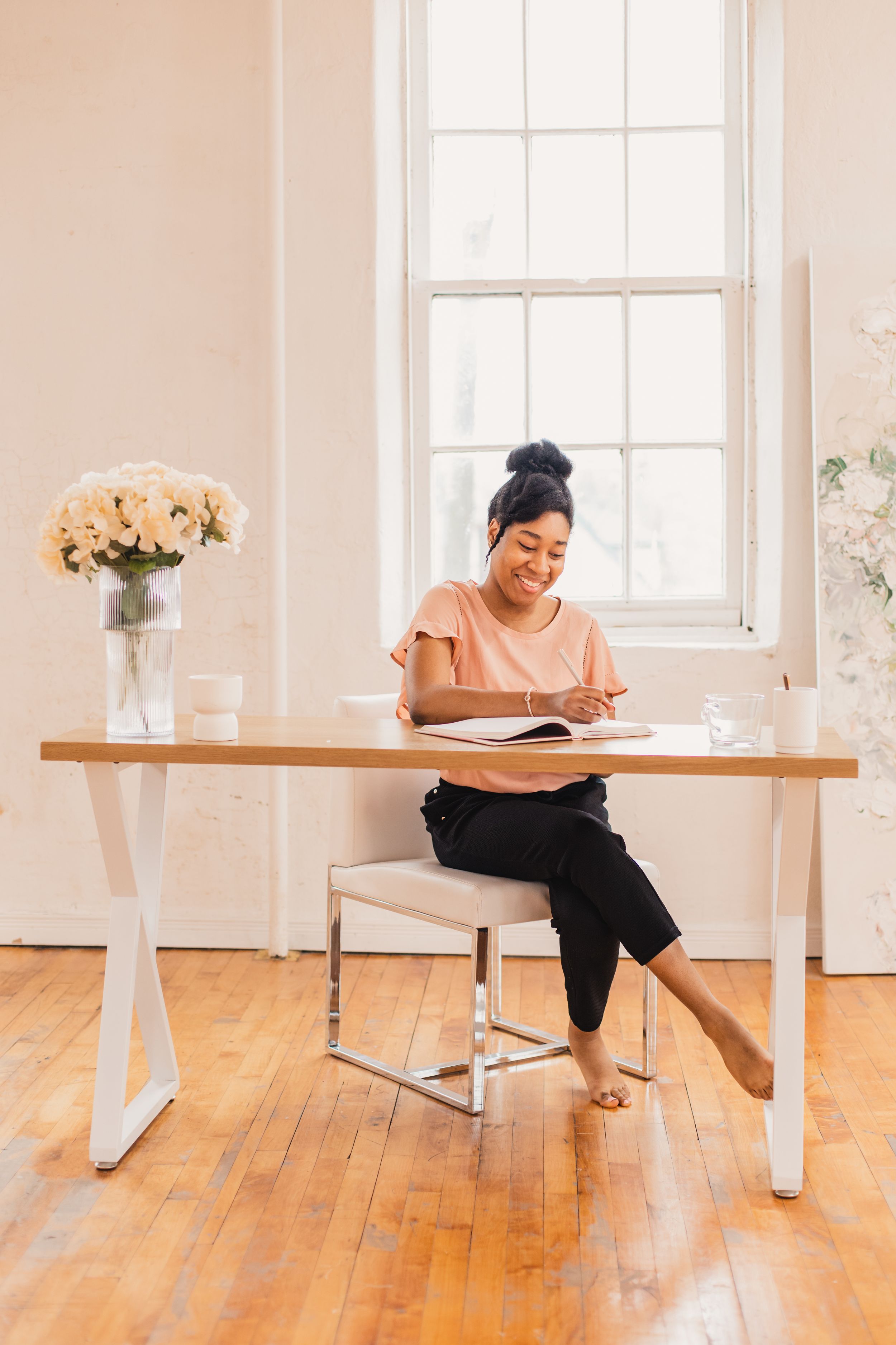 Someone writes at a modern white desk with flowers in a bright room with hardwood floors and natural light.