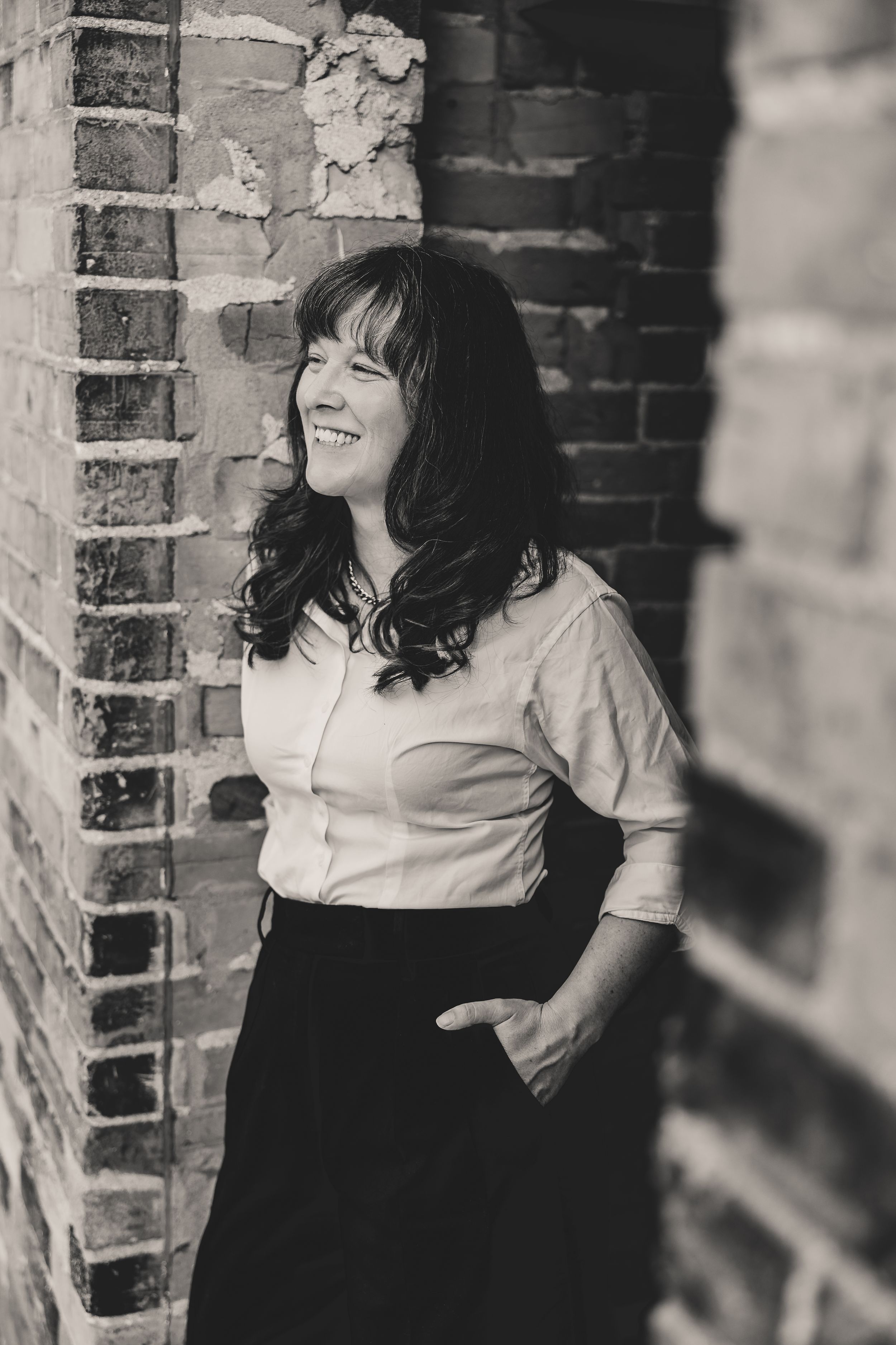 A black and white portrait of someone in a white blouse and dark skirt laughing against a brick wall.