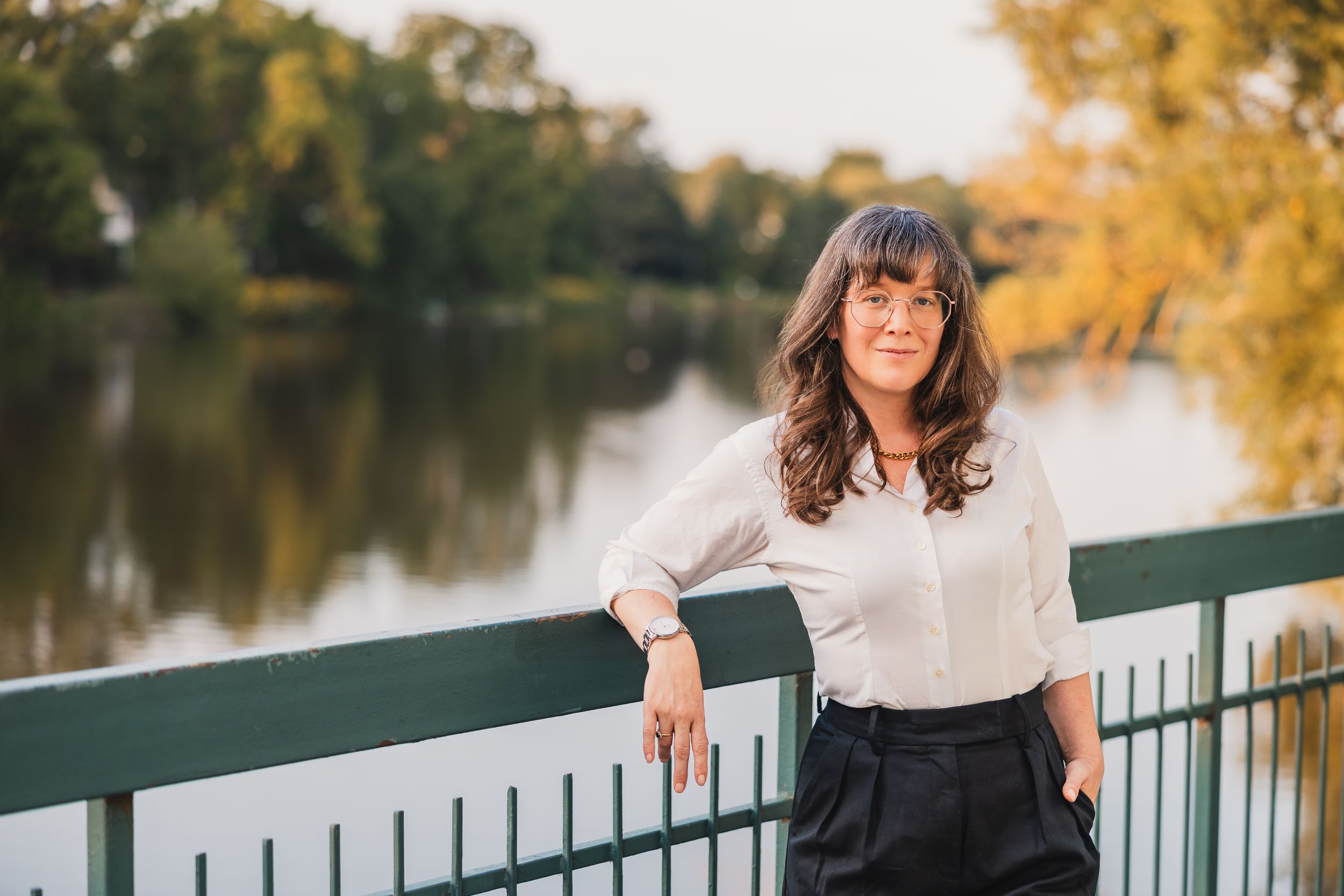 A person in a white blouse and dark skirt stands at a railing overlooking a serene lake during golden hour.