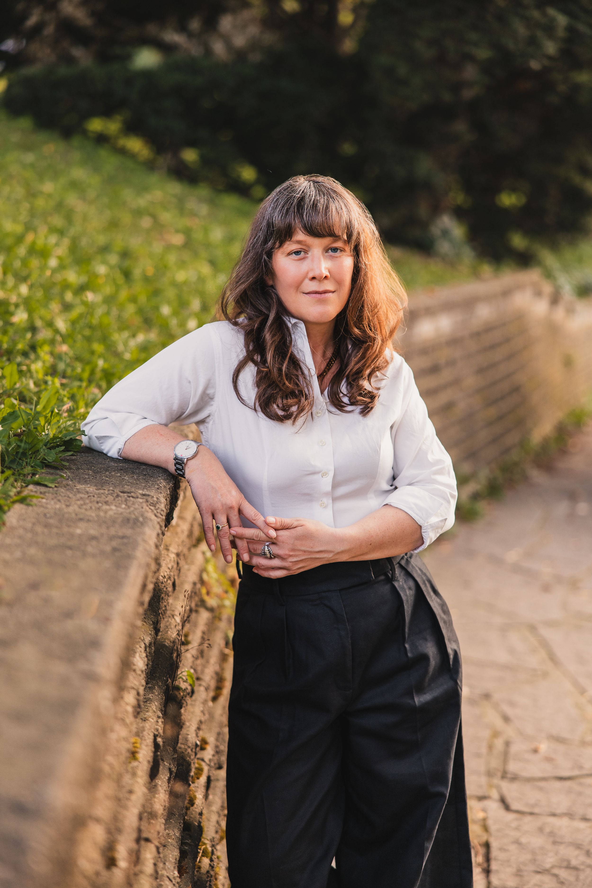 A person in a white button-up shirt and dark pants poses casually outdoors on a stone wall in natural lighting.