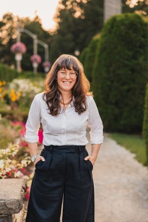 A person in a white button-down shirt and navy pants stands smiling on a garden path at sunset.