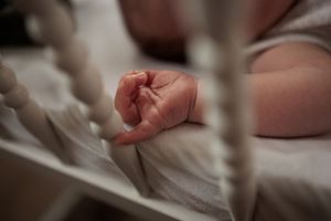 Newborn baby's tiny hand resting on white crib mattress, photographed through white crib slats.