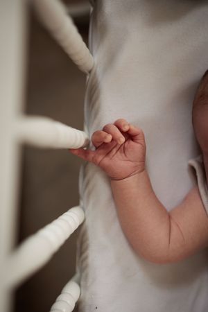 Close-up of a newborn baby's small hand gripping a white crib rail from inside the crib.